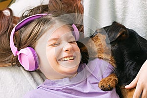 Happy teenage girl in lilac t-shirt in pink headphones listens to music and has fun playing with dog