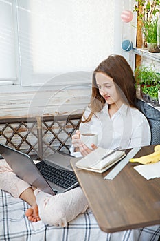 Happy teenage girl sits in her pajamas and learns remotely via the Internet during quarantine. Drinking tea for