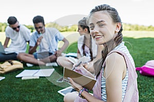 happy teenage girl taking notes and smiling at camera while studying with friends