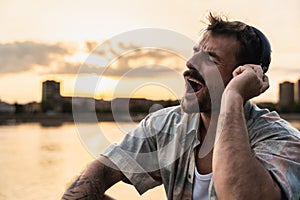 Happy teenage male tourist sitting on sea promenade of an old city in sunset. Young man world traveler singing his favorite song