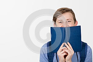Happy teenager boy smiling covering half face with a opened blue book. Book lover. Ready to study hard.
