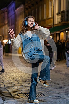 Happy teenager child girl in wireless headphones listening music in smartphone dancing, city street
