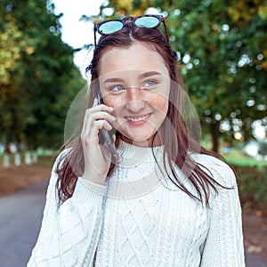 Happy teenager girl smiles and laughs, calls on phone, summer city park, background autumn trees. Emotions happiness are
