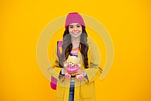 Happy teenager school girl in warm hat, autumn kids portrait. School teenager child girl with backpack hold toy. Fun