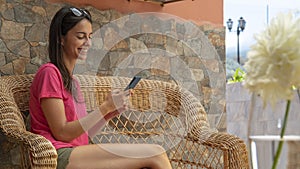 Happy teenager smiling, sending message by cellphone. Good looking girl sitting in wicker couch chatting with friends. Woman