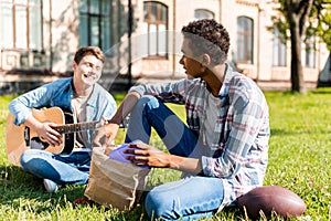 happy teenagers sitting on grass and playing acoustic guitar