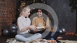 Happy teens exchanging gifts. Two pretty young african and caucasian girls sitting near a New Year or Christmas tree and
