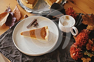 Happy Thanksgiving. Pumpkin pie slice on modern plate and hot coffee on rustic table with linen napkin, autumn flowers and leaves