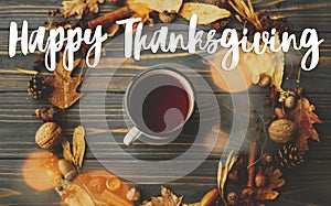 Happy Thanksgiving text sign on metal mug with tea and autumn wreath made of leaves, berries, acorns and pine cones on dark wood.