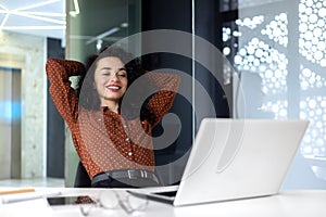 Happy thinking woman at workplace inside office, business woman with hands behind head completed work well, latin