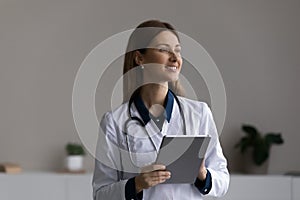 Happy thoughtful female doctor, physician using tablet computer at office