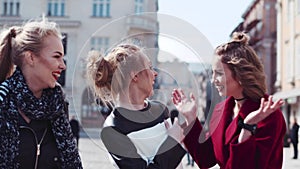 Happy three friends wandering in the old city center, laughing, and joking. The woman in a red coat hugs her friend