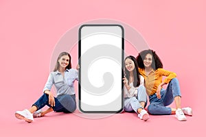 Happy three multiracial female friends sitting near giant cellphone with white blank screen, mockup, free space