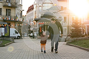 A happy three year old son walks hand in hand with his dad in the city on a sunny spring day