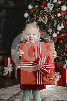 Happy three-years old girl holds large gift box and looks into camera on xmas tree background. Christmas Eve. Vertical frame