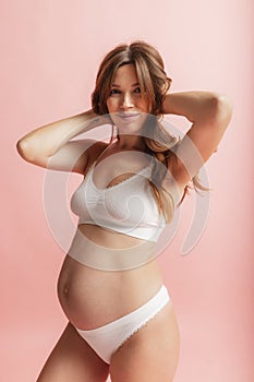 Happy time, awaiting. Portrait of smiling beautiful pregnant woman in lingerie isolated over pink studio background