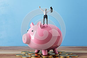 Happy tiny businessman celebrating success while standing on pink piggy bank and golden coins. Blue concrete wall background.