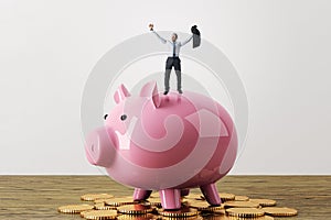 Happy tiny businessman celebrating success while standing on pink piggy bank and golden coins. White concrete wall background.