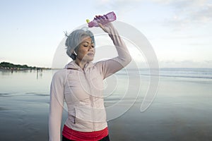 Happy tired and thirsty middle aged woman drinking and pouring water on her head refreshing after beach running workout - 40s or