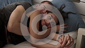 Happy to be together. Indoor portrait of joyful young male and female lying in bed covered with white sheets, smiling