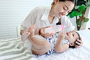 Happy toddle baby infant child lying on white bed while mother holding milk bottle for feeding her daughter kid girl, mom love and