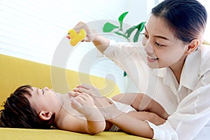 Happy toddle baby lying on yellow sofa in living room, mom holding duck toy for playing with little girl daughter, parent play