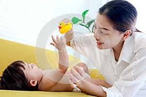 Happy toddle baby lying on yellow sofa in living room, mom holding duck toy for playing with little girl daughter, parent play