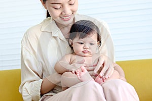 Happy toddle baby sitting on mother lap on yellow sofa in living room at home, mom bonding with little girl daughter, parent photo