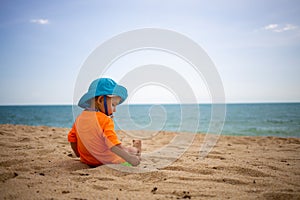 A happy toddler in blue hat and orange t-sirt plays sand on a tropical beach and picking on the sand. sea is on