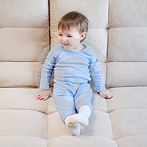 Happy toddler boy in blue clothes sits on a beige sofa. Child age one ye