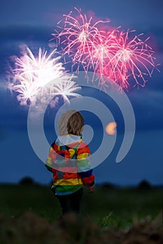 Happy toddler boy, child, looking at fireworks for 4th of July in a fiel, full moon rising up