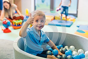 Happy toddler boy playing in a ball pit full of colorful balls, showing thumb up. Sensory play at the nursery school for