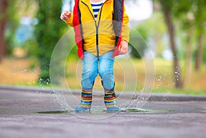 Happy toddler boy with umbrella and color rubber boots jump on a rainy puddle in a park, carefree childhood
