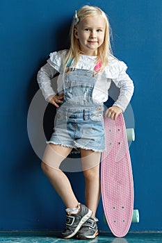 Happy toddler girl with blond hair holding a skateboard while posing on blue wall background