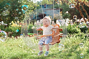 Happy toddler girl having fun on a swing. Sunny summer playground in a kindergarten. Soap bubbles flying around. The