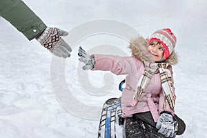 A happy toothless girl is sitting on a sledge in a winter park, reaching for an adultÃ¢â¬â¢s hand before touching photo