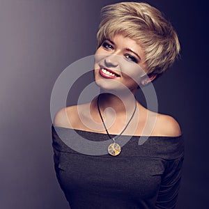 Happy toothy smiling woman with short blond bob hairstyle in grey blouse and orange fashion necklace on gray background. Closeup