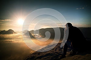 Happy tourist with camera in hands sit on peak of sandstone rock and watching into colorful mist and fog in morning valley.