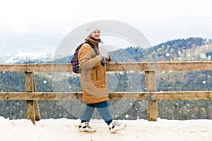 Happy tourist senior woman with backpack standing on observation deck and looking at beautiful mountains covered snow