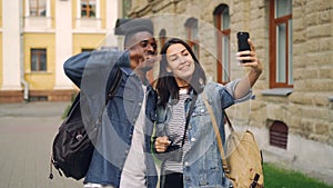 Happy tourists friends with backpacks are taking selfie using smartphone showing hand gestures v-sign and thumbs-up