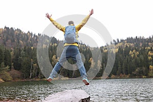 Happy traveler with backpack jumps with open arms, in background lake and mountains.