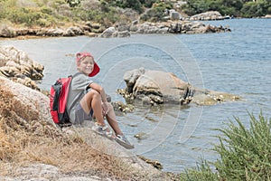 A happy traveler boy with backpack is sitting on rocks near the sea