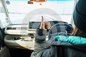 Happy Traveler driving car on snowy road and gesture finger up, woman Tourist enjoying snow forest view from the car in winter