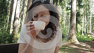 Happy traveler lady blows and drinks hot tea from thermos cup during forest summer hike. Refreshment and relaxing moment