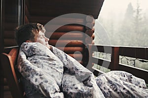 Happy traveler man resting in blanket on wooden porch with view on woods and mountains. Space for text. Hipster relaxing in