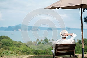 Happy traveler in white shirt and hat enjoy beautiful sea view, young man sitting on chair and looking ocean at tropical beach.