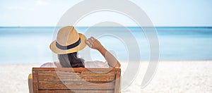 Happy traveler woman in white dress and hat enjoy beautiful sea view, young woman sitting on chair and looking ocean at tropical