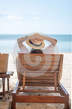 Happy traveler woman in white dress and hat enjoy beautiful sea view, young woman sitting on chair and looking ocean at tropical