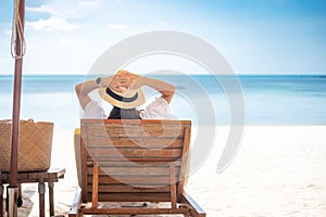 Happy traveler woman in white dress and hat enjoy beautiful sea view, young woman sitting on chair and looking ocean at tropical