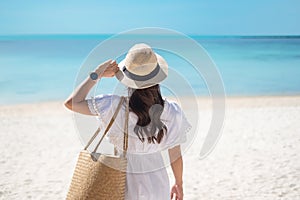 Happy traveler woman in white dress and hat enjoy beautiful sea view, young woman standing on sand and looking ocean at tropical